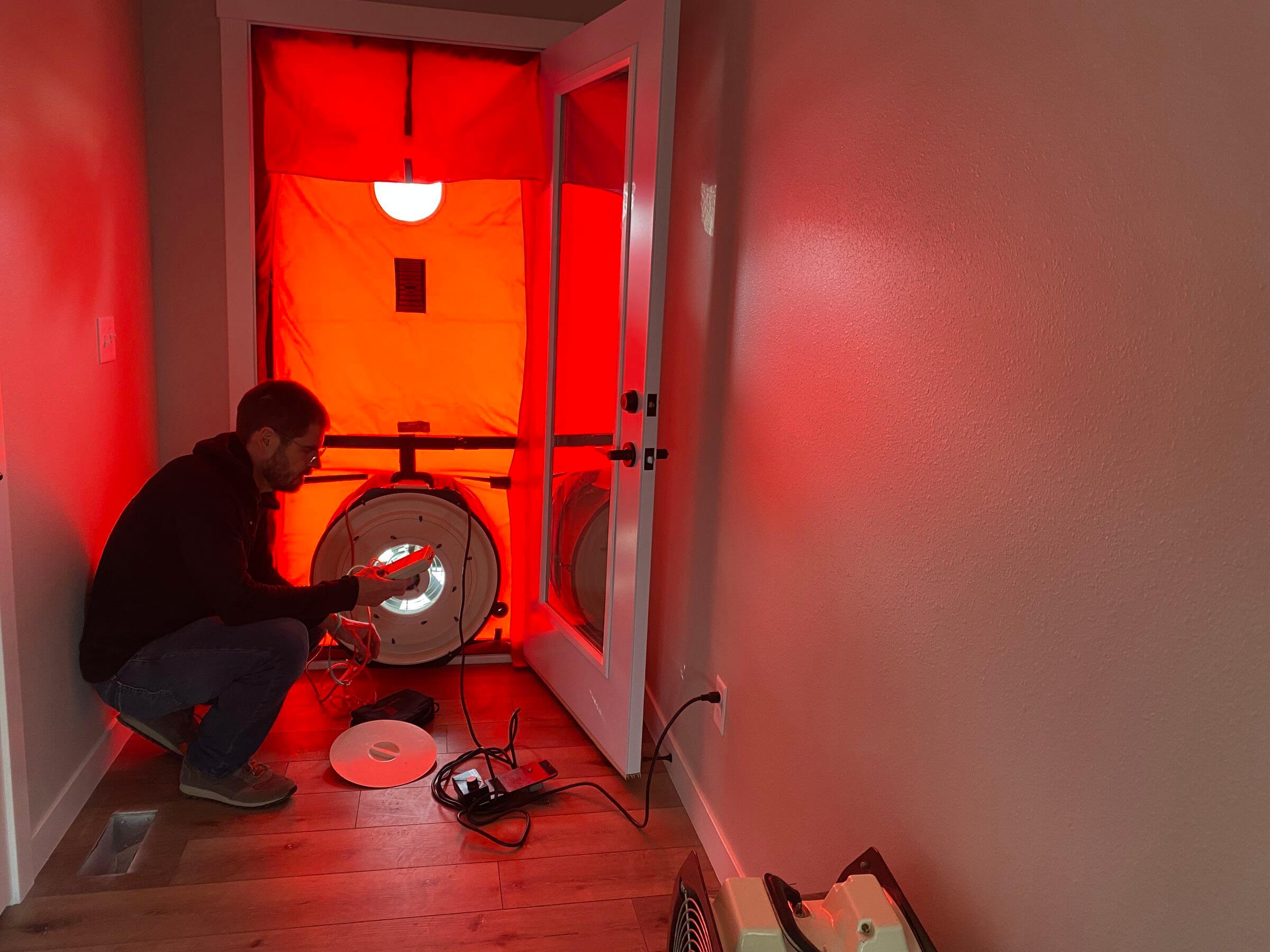 A person sets up a blower door test in a hallway, using a red fabric panel and fan system to check for air leaks in a building.