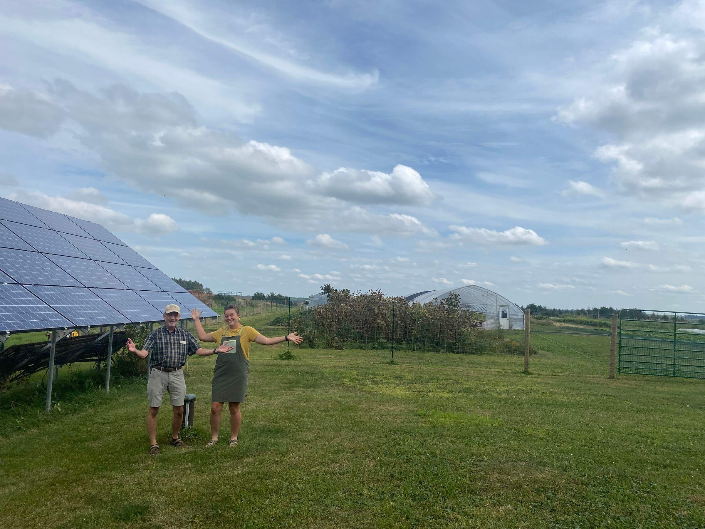 Two people stand on grass near solar panels, with greenhouses and a fenced area in the background under a partly cloudy sky.