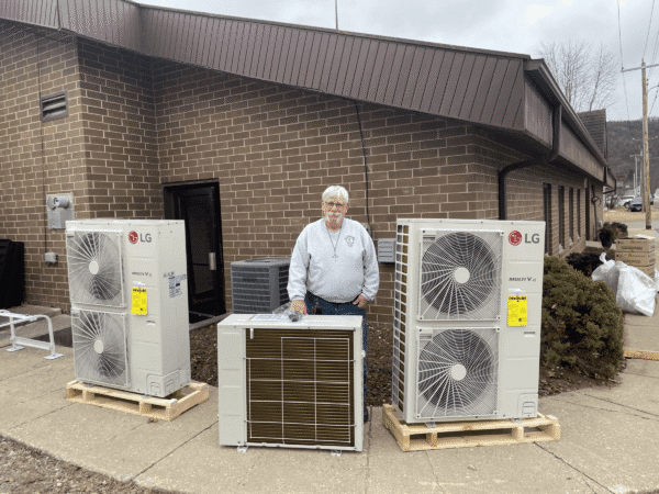 A man stands outside a brick building behind three large LG air conditioning units placed on wooden pallets.