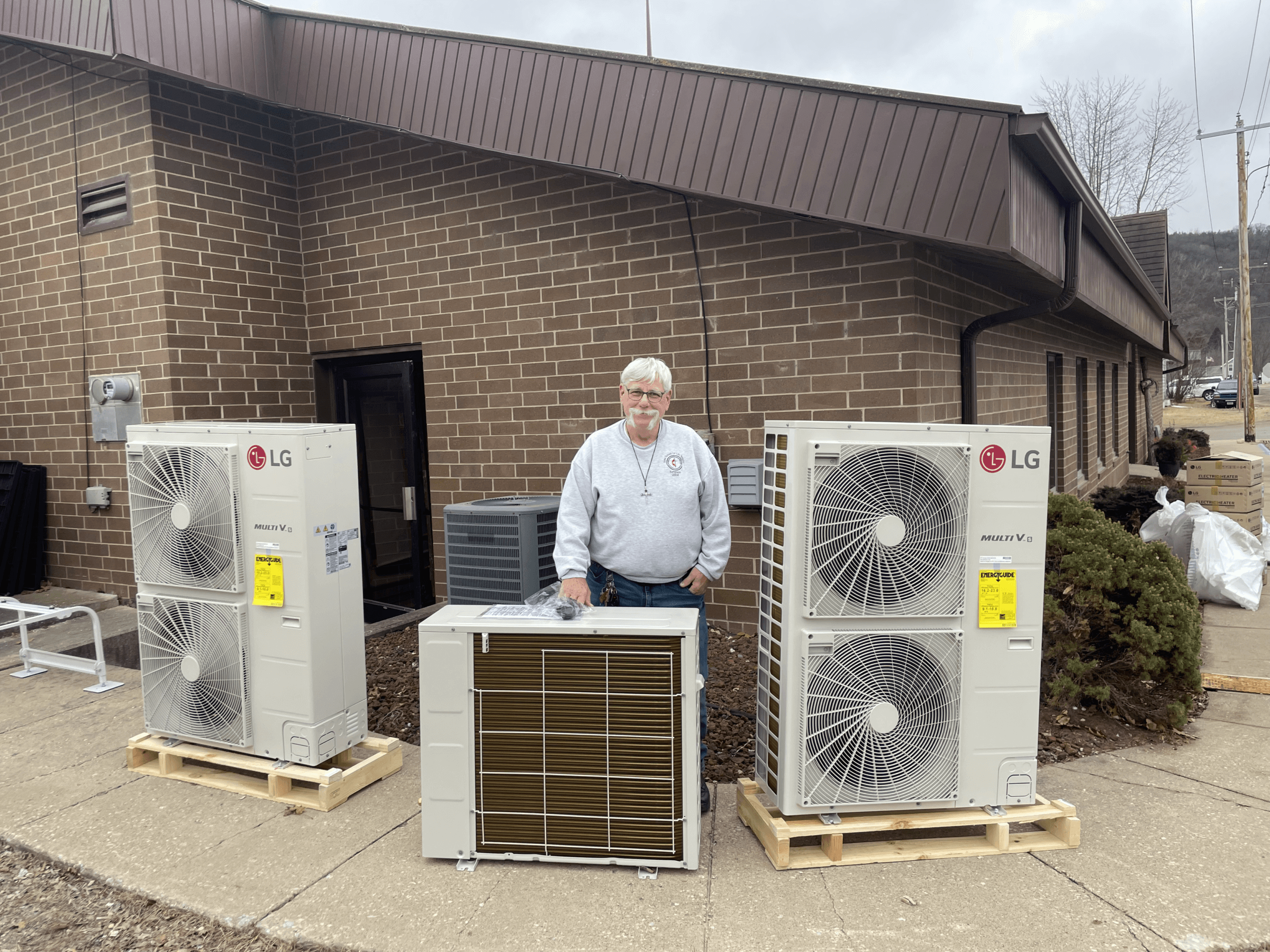A man stands outside a brick building behind three large LG air conditioning units placed on wooden pallets.