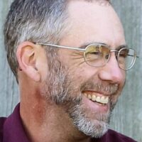 A middle-aged man with glasses and a graying beard smiles while looking to the side, standing in front of a wooden background.