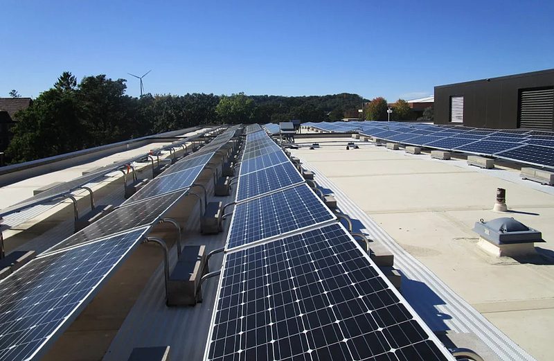 Rows of solar panels are installed on the flat rooftop of a building under a clear blue sky, surrounded by trees and distant buildings.