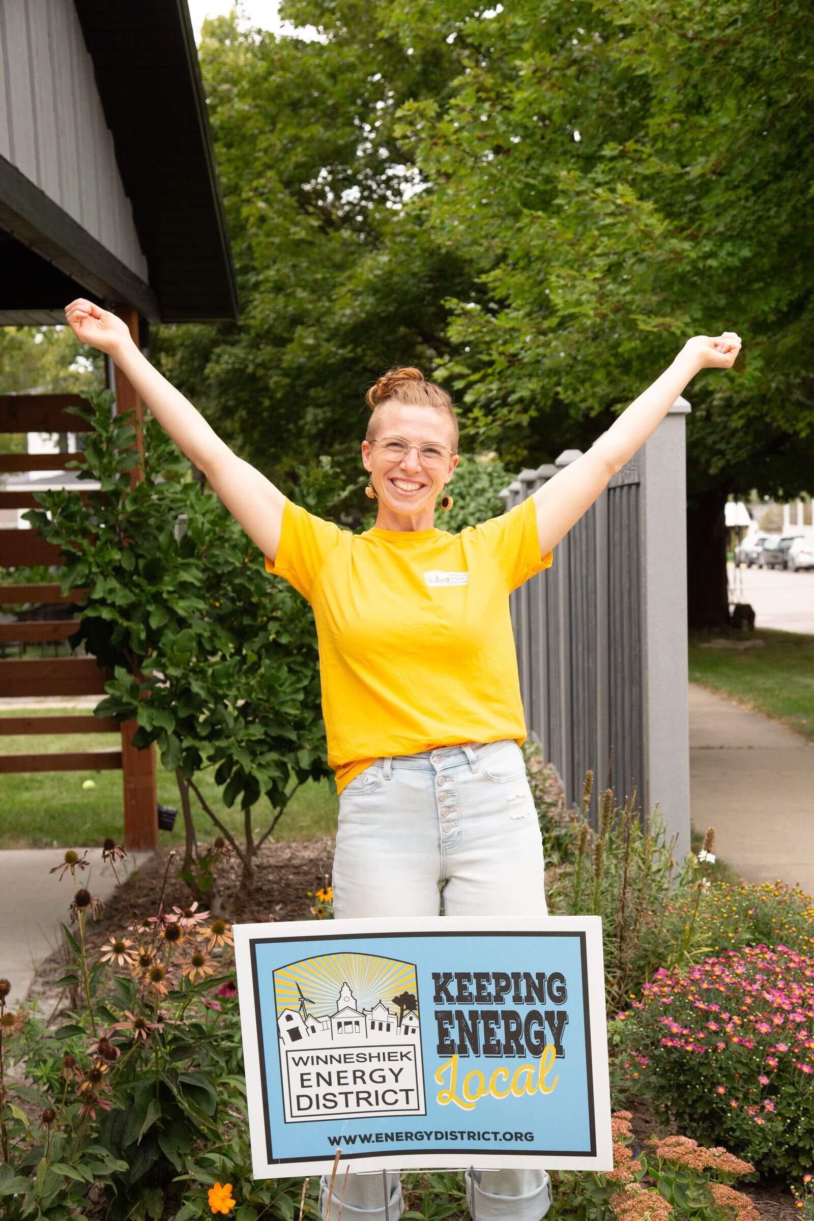 A person in a yellow shirt stands outside with arms raised, smiling behind a “Winneshiek Energy District: Keeping Energy Local” sign.