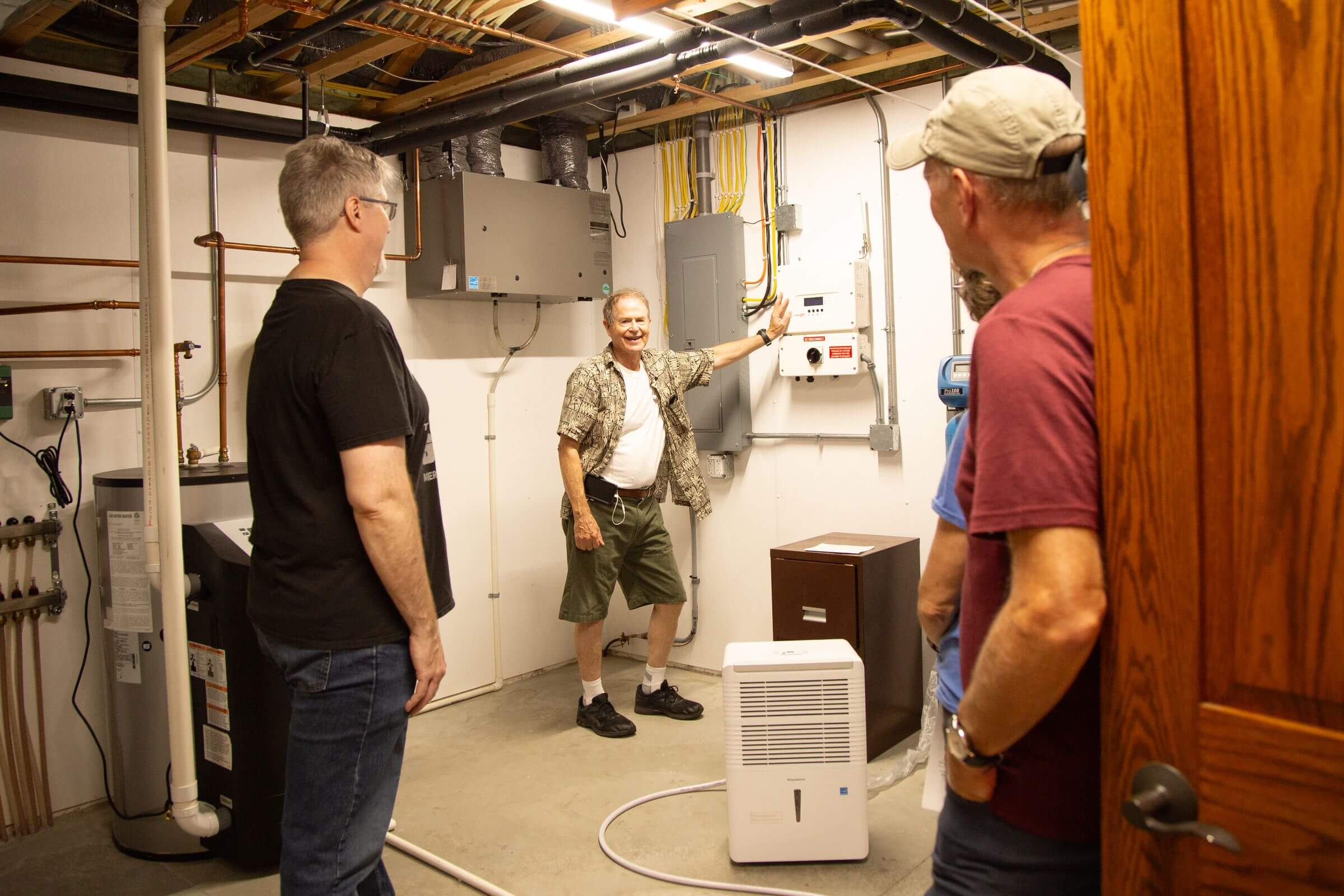 Four men stand in a utility room with exposed pipes and electrical panels; one man points at a panel while others observe.