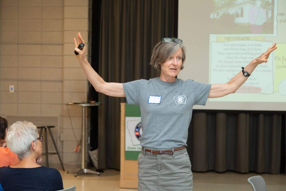 A woman stands with arms raised, speaking to an audience in a room with a presentation screen and podium in the background.