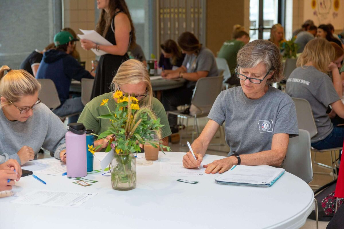 Several people sit around a round table, filling out paperwork with pens. A vase with yellow flowers is in the center of the table. More people are in the background.