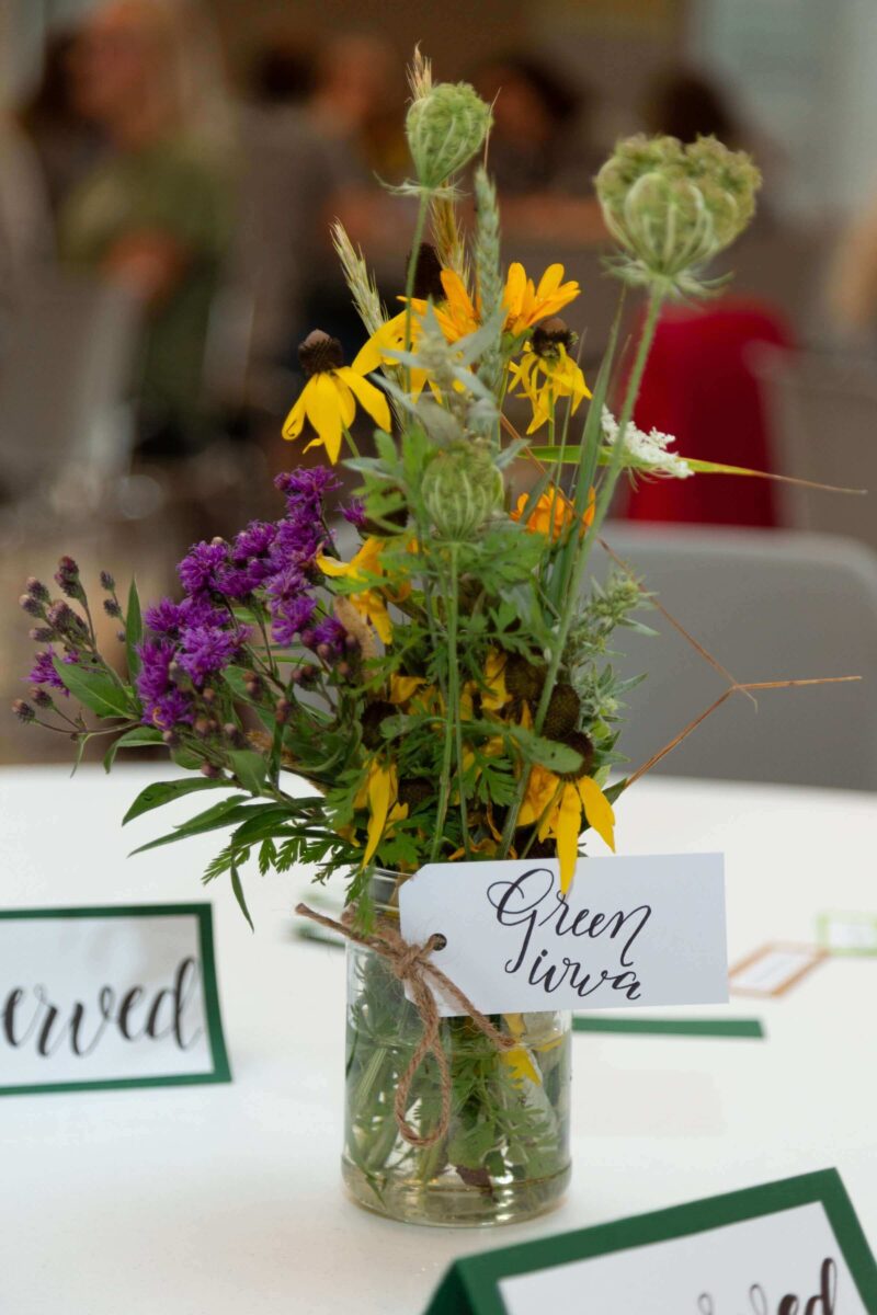 A small glass jar filled with assorted wildflowers sits on a table, labeled 