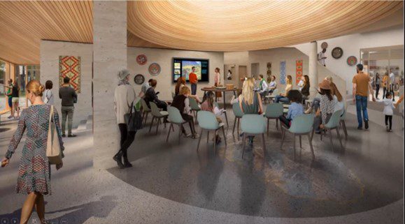 People sit and stand in a spacious, modern room with a circular ceiling, watching a presentation on a screen; artworks and decor are displayed on the walls.