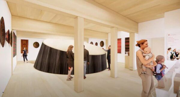 People explore an art gallery with wooden floors and columns; a woman holds two babies in the foreground, and a large circular installation is in the center of the room.