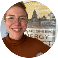 Person with short hair and glasses smiles in front of a sign that reads “INNESHEK ENERGY PROJECT” with a building silhouette and sunburst in the background.