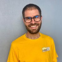 A man with short brown hair, a beard, and glasses is smiling. He is wearing a bright yellow T-shirt with a small logo on the chest and standing in front of a plain gray background.
