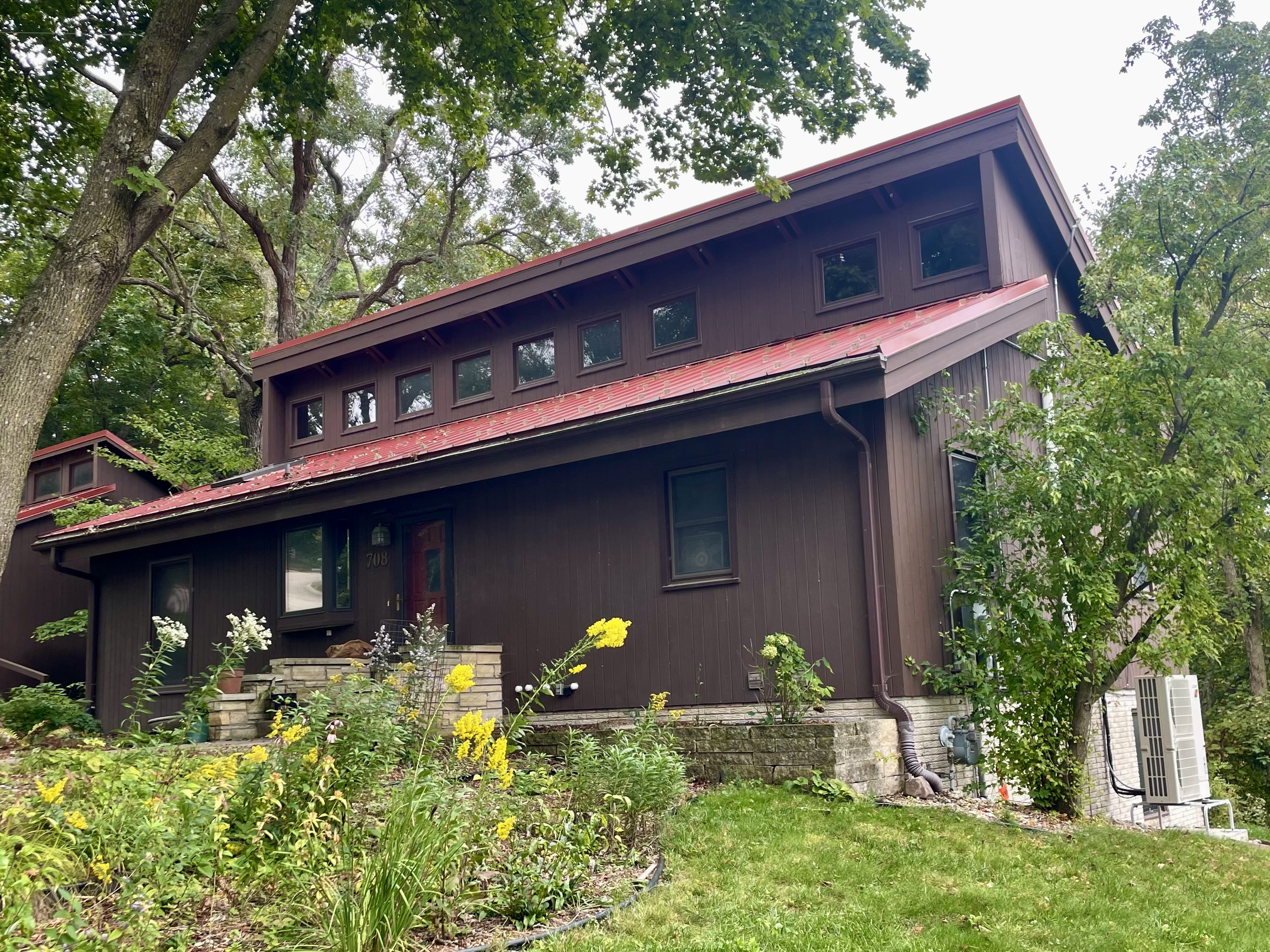 A mid-century house, brown with red metal roof, surrounded by trees.